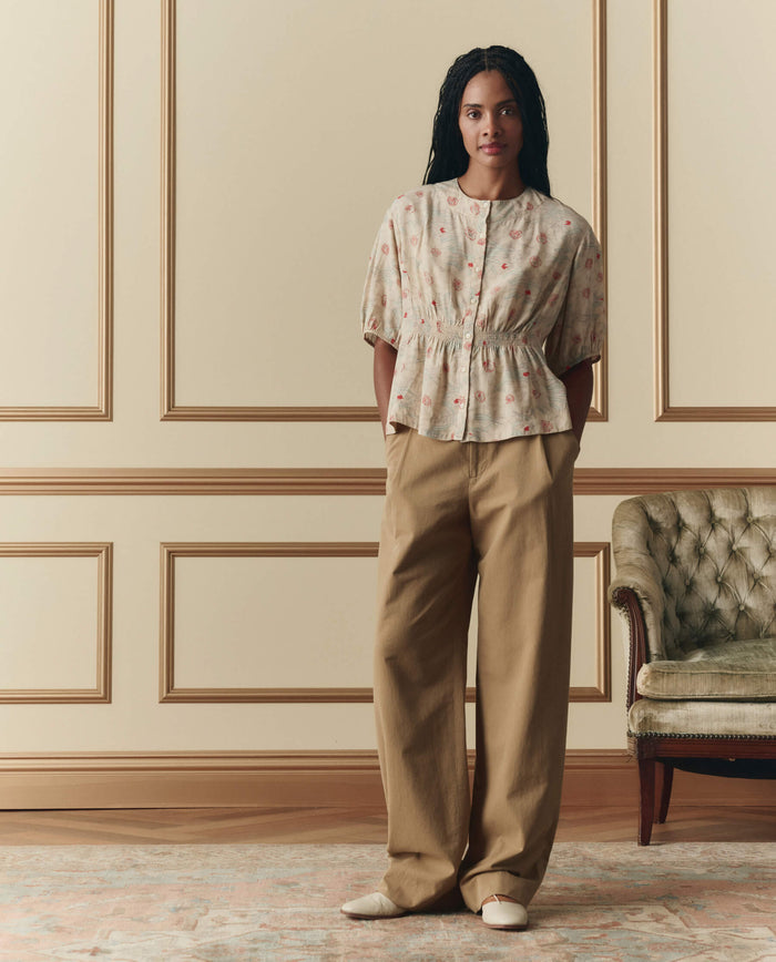 A woman stands indoors by a green tufted sofa, hands in her pockets, wearing THE GREAT. SP26 D2's The Hayflower Top in Champagne Scattered Bloom, tan wide-leg pants, and cream shoes.