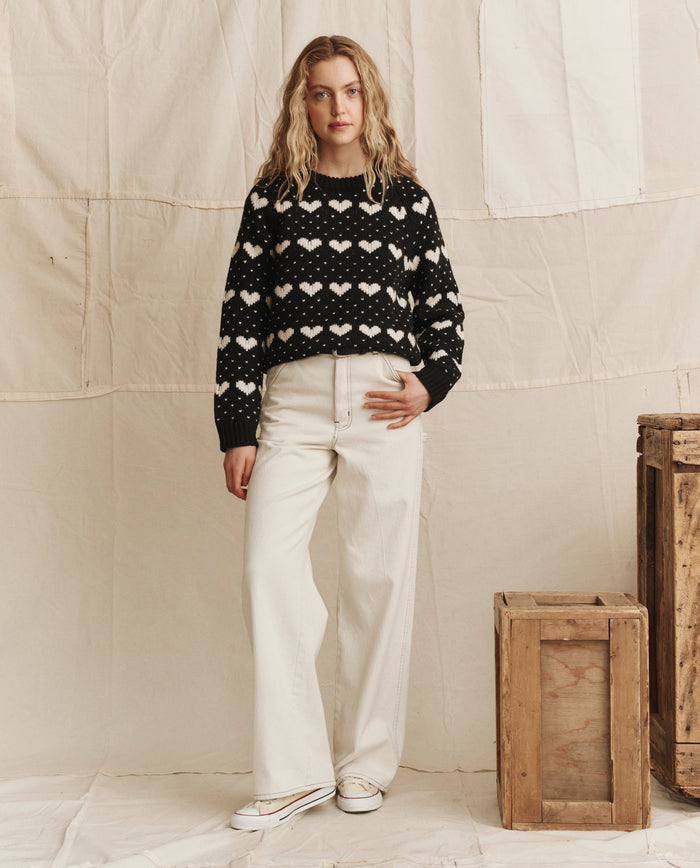 A woman stands indoors next to wooden crates against a neutral backdrop, wearing THE GREAT. PS26's The Heart Crew. in Black With Pearl, off-white wide-leg pants, and white sneakers.