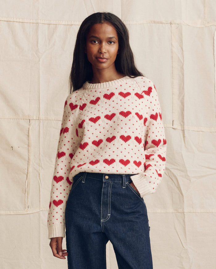 A woman stands before a beige fabric backdrop wearing THE GREAT. PS26's The Heart Crew. sweater in pearl with red and dark blue high-waisted jeans.