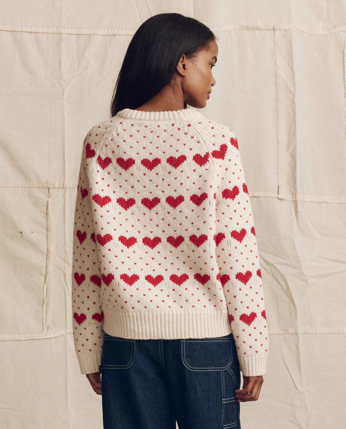A woman faces away against a neutral fabric backdrop, wearing THE GREAT. PS26's The Heart Crew. sweater in Pearl with Red hearts and blue jeans.