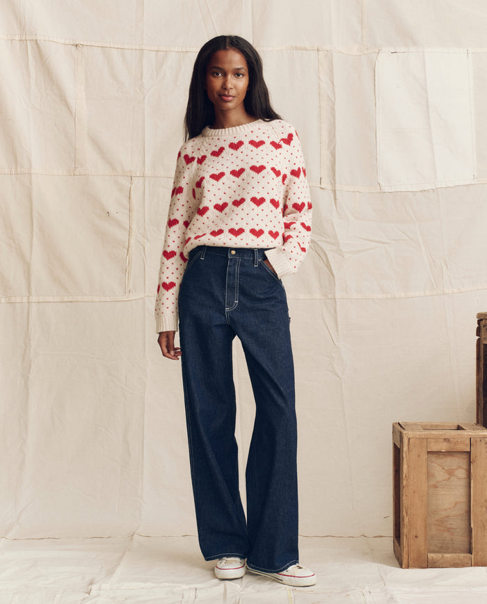 A woman stands against a neutral fabric backdrop wearing THE GREAT. PS26 The Heart Crew. sweater in Pearl with Red, paired with dark wide-leg jeans and white sneakers; wooden crates sit to the right.