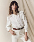 A person with long hair models The Hemingway Top by THE GREAT. PS25, paired with stylish pants against a neutral background, hand resting on hip.