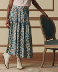 A person wearing a white top, beige lace-up boots, and THE GREAT. SP26 D2 The Highland Skirt – Blue Scattered Bloom stands beside a green upholstered wooden chair.