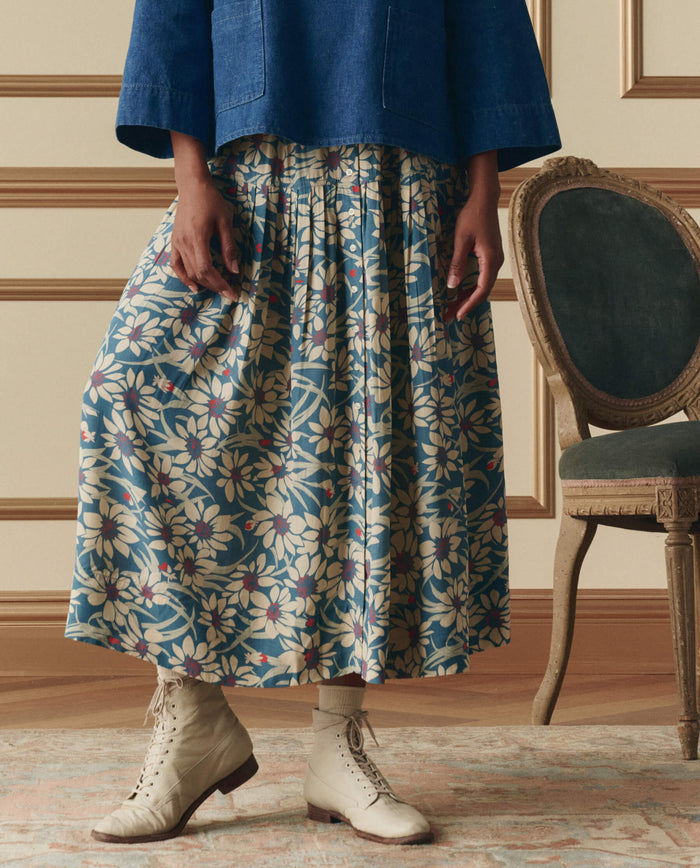 Indoors, a person in a blue top and THE GREAT. SP26 D2 The Highland Skirt – Blue Scattered Bloom stands by a vintage green upholstered chair, wearing white lace-up boots.