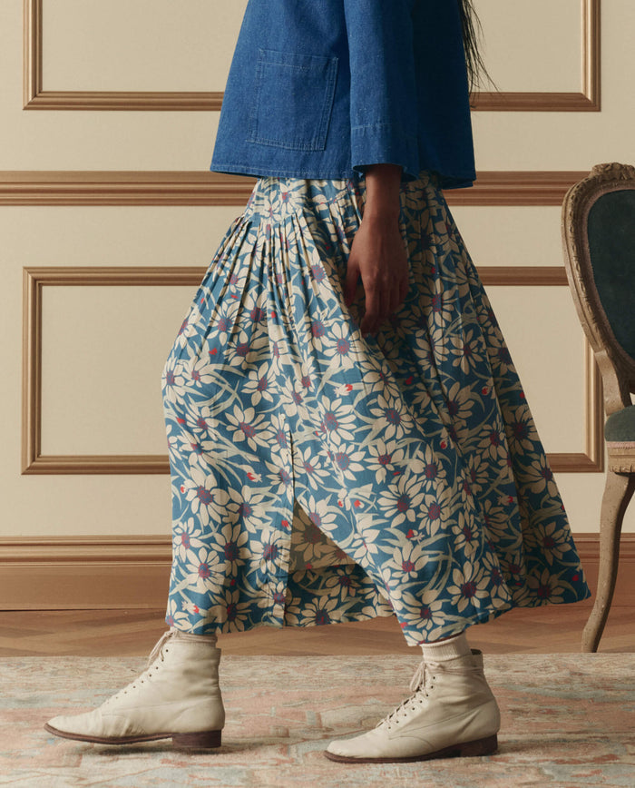 Wearing THE GREAT. SP26 D2’s The Highland Skirt in Blue Scattered Bloom, a person stands on a rug in a blue jacket, white lace-up boots, and socks, with a vintage chair visible in the background.