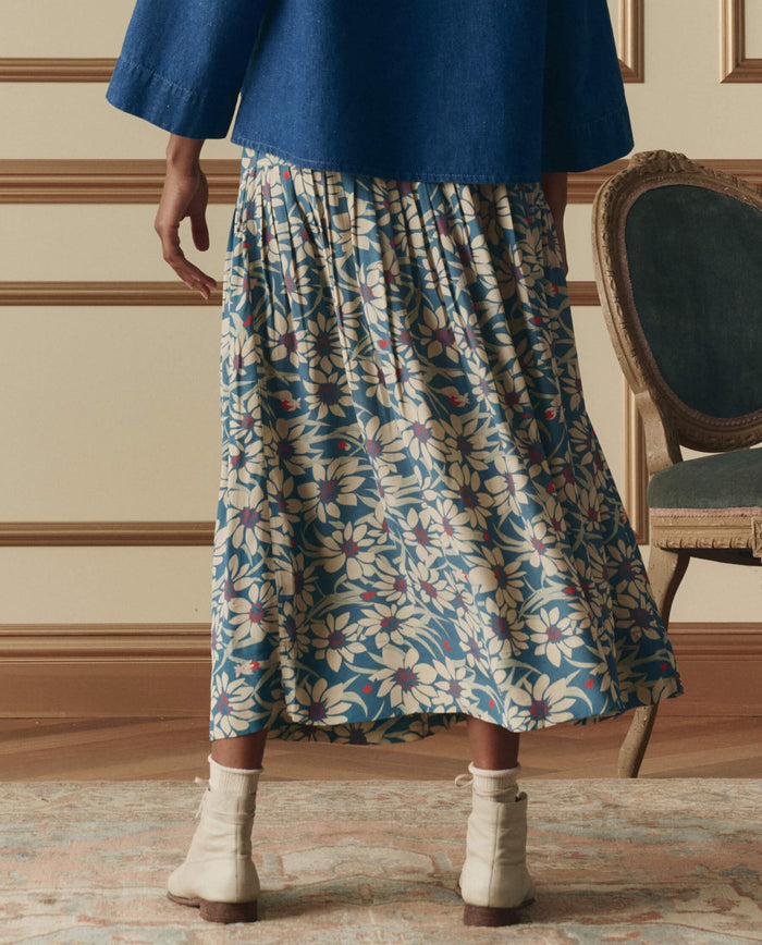 Indoors, a person wears THE GREAT. SP26 D2's The Highland Skirt in Blue Scattered Bloom with light boots and a blue top. A vintage chair and patterned rug enhance the setting’s charm.