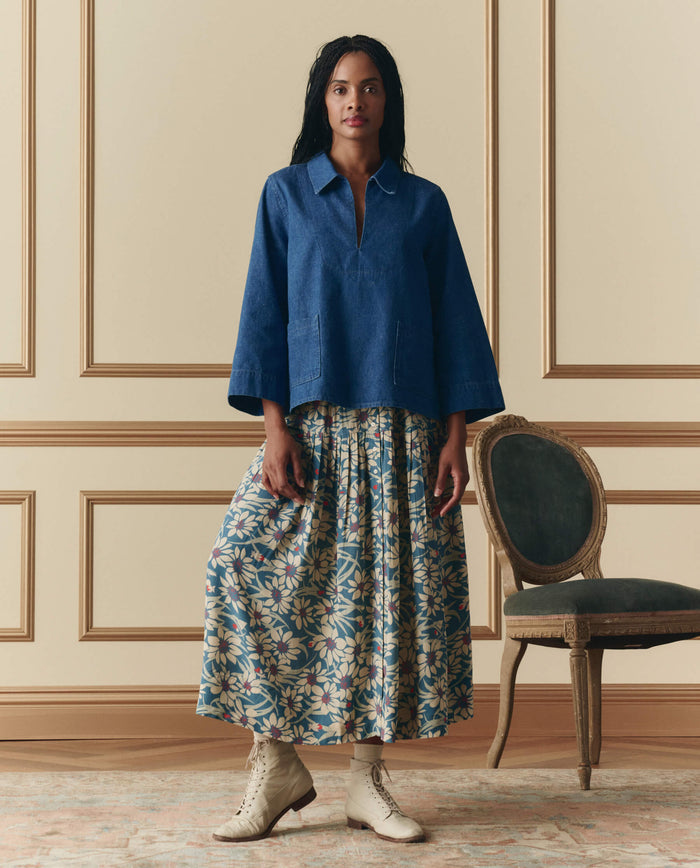 A woman stands indoors beside an empty green chair, wearing THE GREAT. SP26 D2’s The Highland Skirt in Blue Scattered Bloom, a blue long-sleeve top, and lace-up boots.