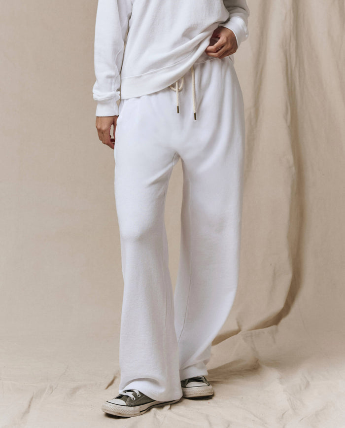 Wearing SP26 New Knits’ The Horseshoe Sweatpant in Solid -- True White with a white long-sleeve top and black-and-white sneakers, a person stands against a beige fabric backdrop, showcasing a relaxed loungewear look.