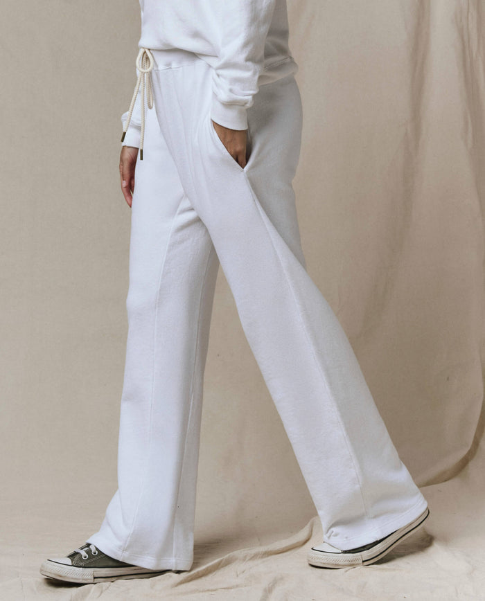 Wearing SP26 New Knits' The Horseshoe Sweatpant in True White, a matching sweatshirt, and black-and-white sneakers, a person stands against a beige fabric background.