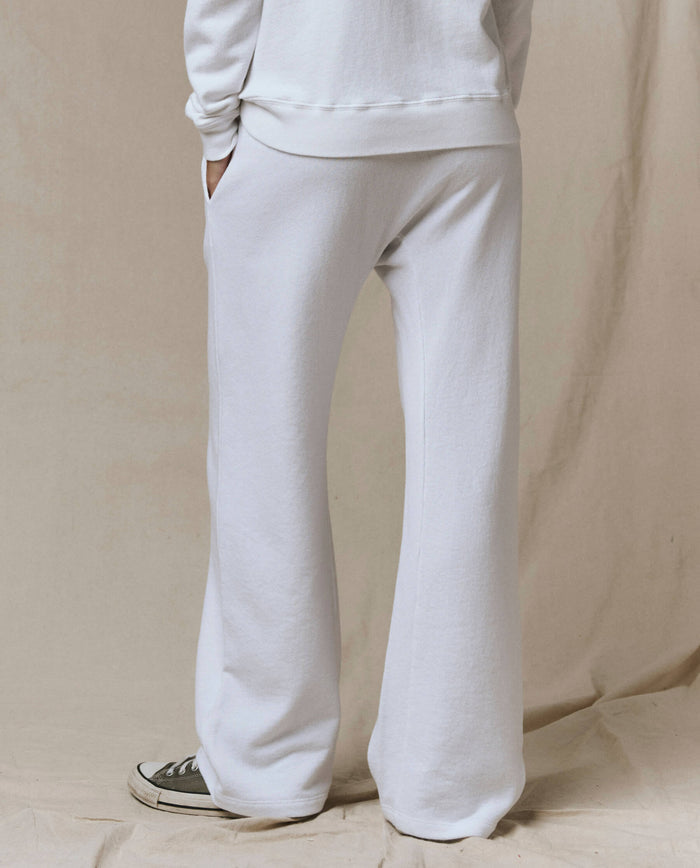 A person stands with hands in pockets, wearing SP26 New Knits' The Horseshoe Sweatpant in Solid -- True White, a matching white sweatshirt, and black-and-white sneakers against a neutral fabric background.