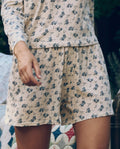 A person wearing The Great. SP26 Sleep's Jersey Boxer Short in Indigo Rose stands beside a patterned quilt.