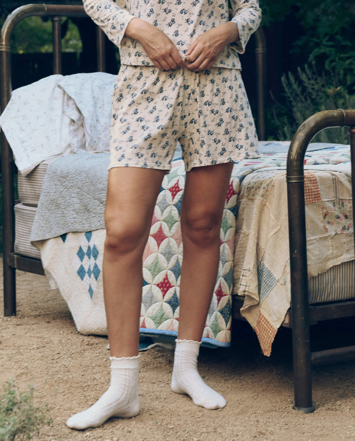 A person wears The Jersey Boxer Short in Indigo Rose by The Great. SP26 Sleep while standing outdoors beside a metal-framed bed layered with quilts and blankets.