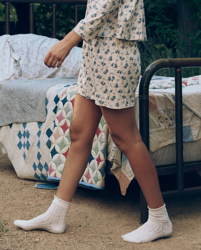 Someone wearing The Great. SP26 Sleep’s Jersey Boxer Short in Indigo Rose walks beside a metal-framed bed with patchwork quilts outdoors, paired with a floral pajama top and white socks.