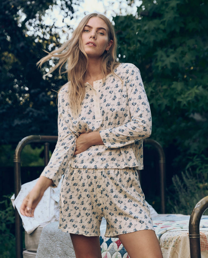 A woman with long blonde hair wears The Great. SP26 Sleep Jersey Boxer Short in Indigo Rose, standing outdoors beside a bed with a quilt.