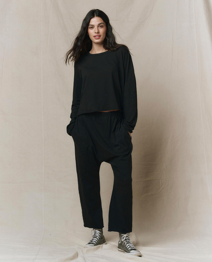 A person stands against a beige backdrop, wearing THE GREAT. CORE KNITS The Long Sleeve Crop Tee in Almost Black, paired with matching black pants and striped sneakers.