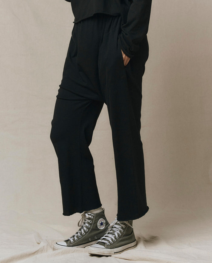 Person stands on a beige surface wearing THE GREAT. CORE KNITS The Jersey Crop Pant in Almost Black, styled with a black long-sleeve top and gray high-top Converse sneakers. Upper body is partially out of frame.