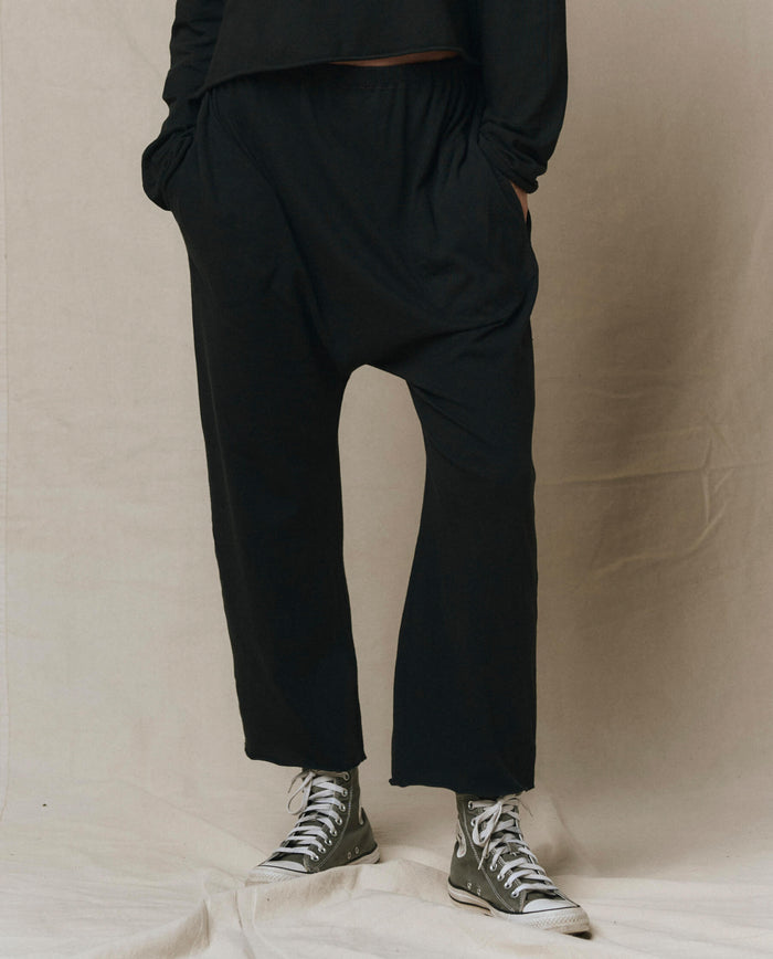 Wearing THE GREAT. CORE KNITS The Jersey Crop Pant in Almost Black, paired with a black long-sleeve top and gray high-top sneakers, a person stands hands-in-pockets against a neutral background.