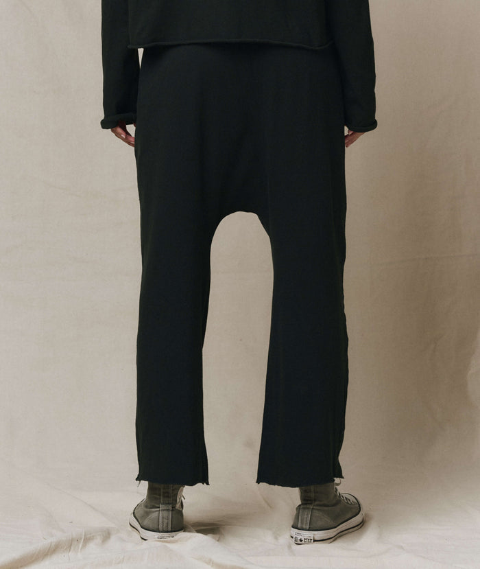 A person stands facing away, wearing THE GREAT. CORE KNITS The Jersey Crop Pant in Almost Black with a dropped rise and elastic waist, styled with long sleeves and gray sneakers against a neutral background.