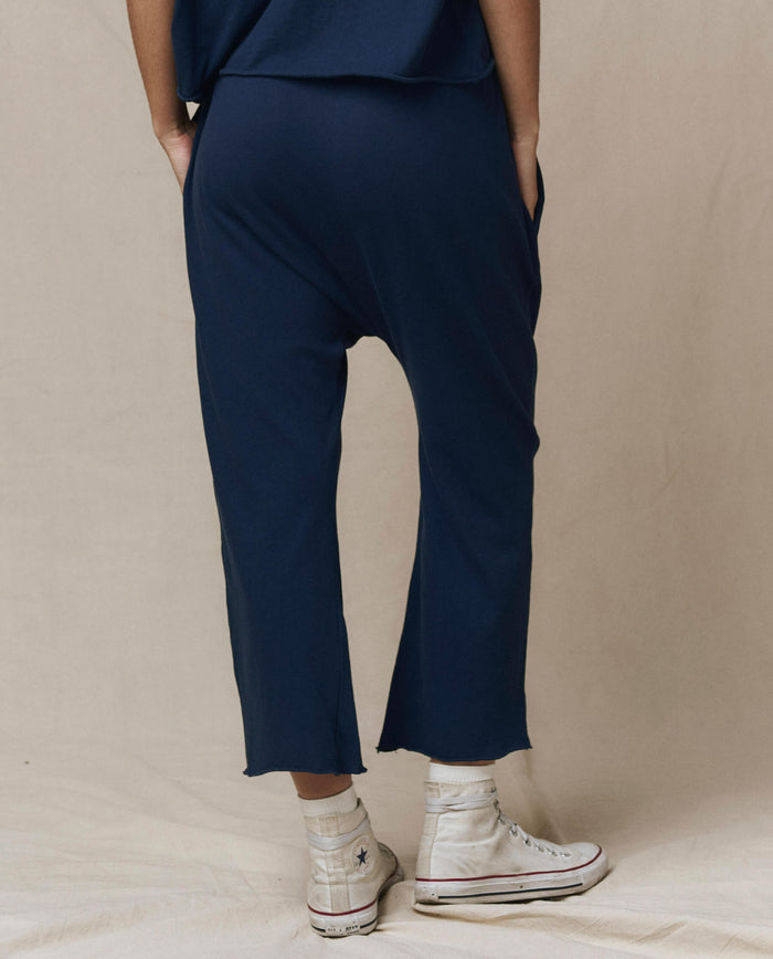 Person wearing THE GREAT. CORE KNITS The Jersey Crop Pant in True Navy with a relaxed fit and elastic waist, paired with white high-top sneakers, stands hands-in-pockets facing away from the camera against a neutral backdrop. Made in the USA.