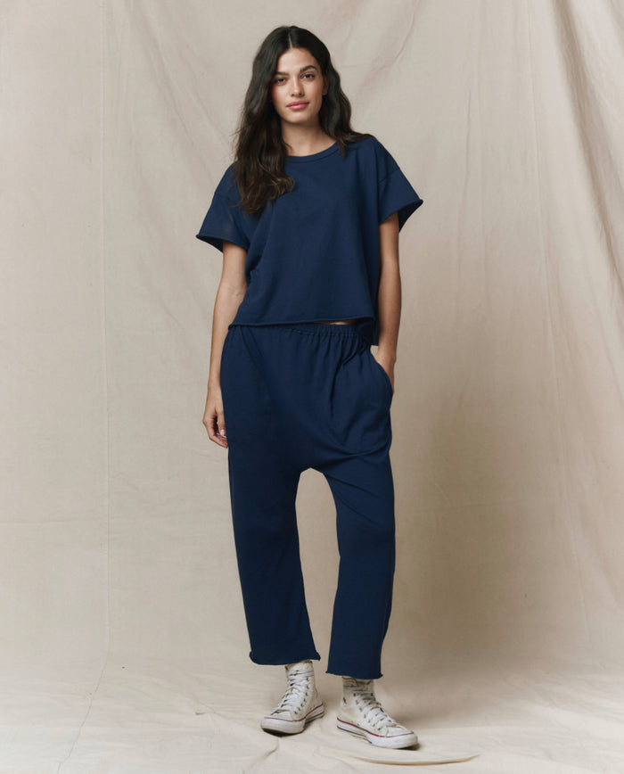 A person stands against a beige backdrop wearing THE GREAT. CORE KNITS' The Jersey Crop Pant in True Navy, paired with a loose-fitting navy top and white high-top sneakers.