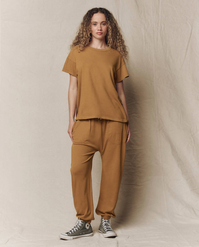 A person stands against a neutral fabric backdrop wearing THE GREAT. SU26 SEASONAL CORE The Jersey Jogger Pant in Cork, a loose brown t-shirt, and gray high-top sneakers.