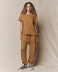 A person stands against a neutral fabric backdrop wearing THE GREAT. SU26 SEASONAL CORE The Jersey Jogger Pant in Cork, a loose brown t-shirt, and gray high-top sneakers.
