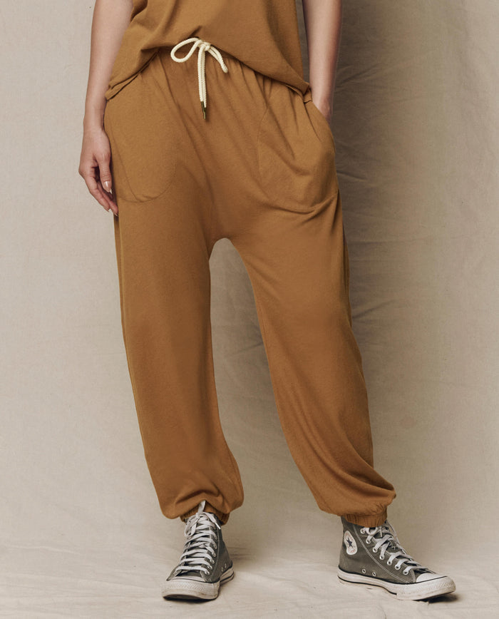 Model wears THE GREAT. SU26 SEASONAL CORE The Jersey Jogger Pant in Cork—loose fit with a drawstring waist and pockets—paired with gray high-top sneakers, standing against a neutral background.