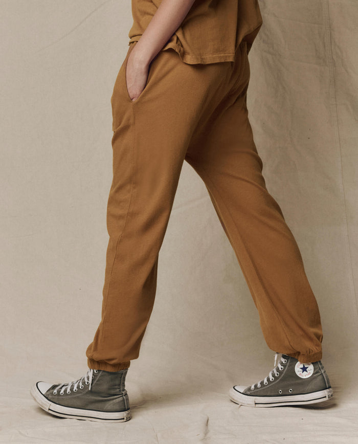 Person wearing THE GREAT. SU26 SEASONAL CORE The Jersey Jogger Pant in Cork, paired with gray high-top Converse sneakers, walking with one hand in their pocket against a neutral background.