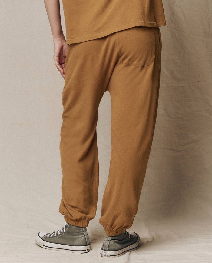 A person stands on a light surface, photographed from the back, wearing THE GREAT. SU26 SEASONAL CORE The Jersey Jogger Pant in Cork and a matching tan top, paired with gray high-top sneakers.