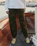 A person stands on a wooden boat deck wearing a white long-sleeve shirt, tan shoes, and THE GREAT. SU26 The Jersey Jogger Pant in Deep Woods Camo.