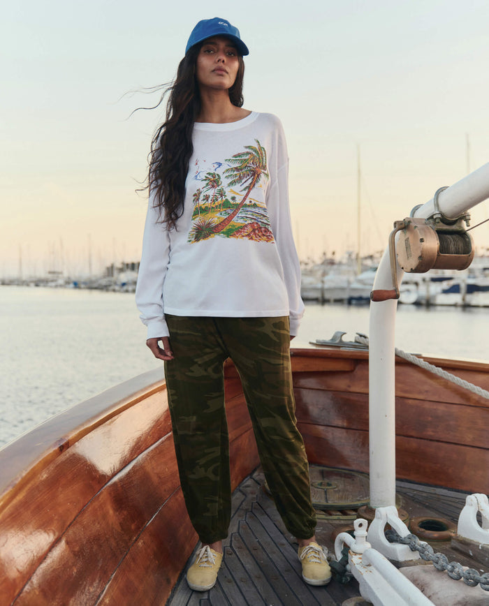A woman stands on a wooden boat in THE GREAT. SU26 The Jersey Jogger Pant in Deep Woods Camo, blue cap, white long-sleeve shirt with a colorful graphic, and tan sneakers. Marina and boats are visible in the background.