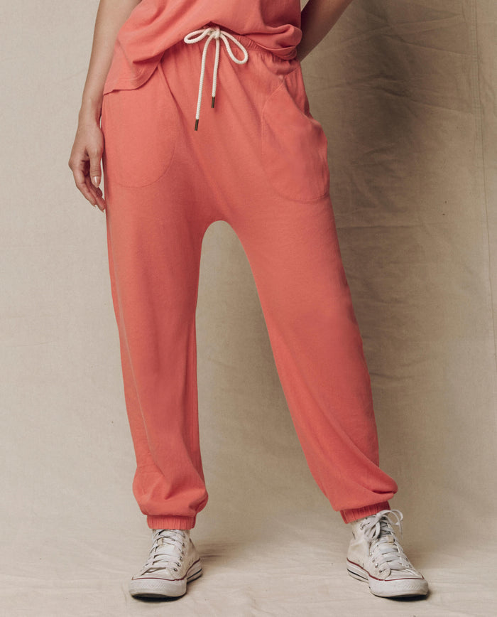 A person wears THE GREAT. SU26 SEASONAL CORE The Jersey Jogger Pant in Guava, featuring an elastic waist and ankle cuffs, paired with a matching top and white sneakers against a plain background.