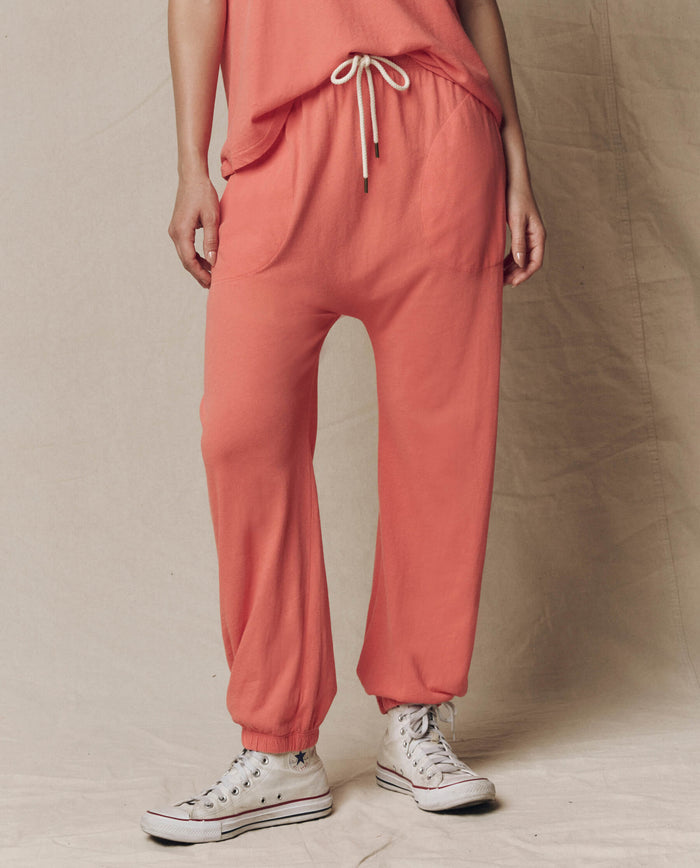 A person wears THE GREAT. SU26 SEASONAL CORE The Jersey Jogger Pant in Guava with a matching top and white sneakers, standing against a neutral fabric backdrop.