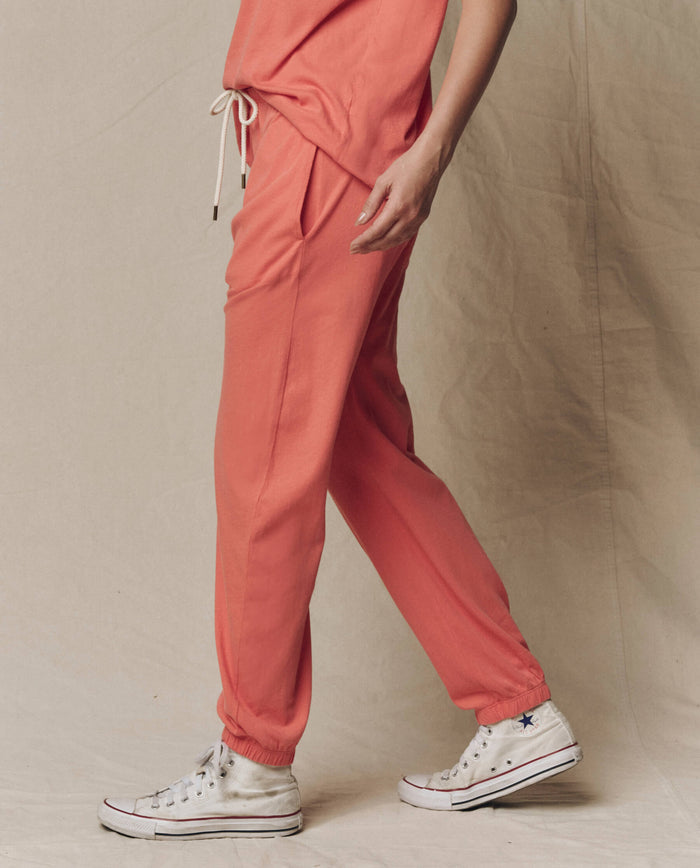 A person stands on a beige background wearing THE GREAT. SU26 SEASONAL CORE The Jersey Jogger Pant in Guava with a matching top and white high-top Converse sneakers.