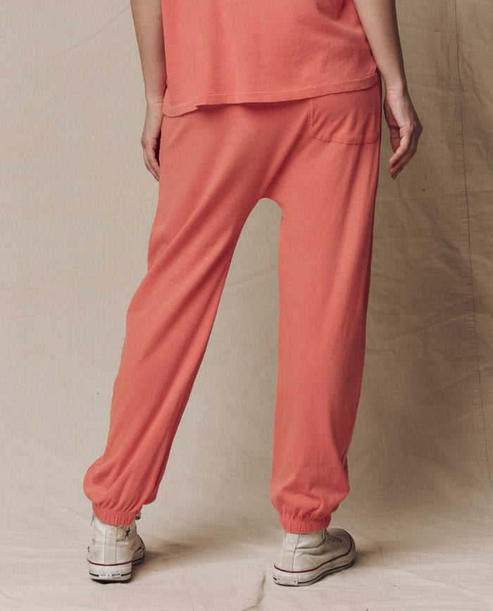 A person faces away from the camera, wearing THE GREAT.'s The Jersey Jogger Pant in Guava (SU26 SEASONAL CORE) and white sneakers, standing against a neutral fabric backdrop.