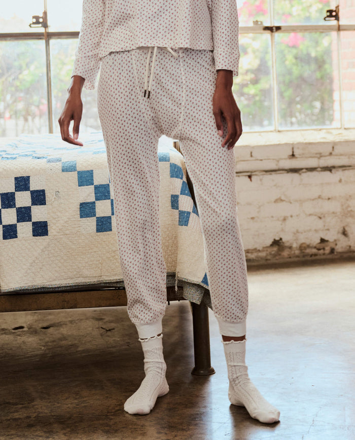 A person stands indoors by a bed, wearing THE GREAT. PS26 SLEEP The Jersey Long John in Calico Rose, with socks on and natural light streaming through a nearby window.