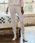 A person stands indoors by a bed, wearing THE GREAT. PS26 SLEEP The Jersey Long John in Calico Rose, with socks on and natural light streaming through a nearby window.