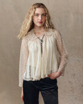 A woman with long wavy blonde hair wears THE GREAT. H25 COLLECTION's The Keepsake Lace Collared Top in cream, styled with a bolo tie and black pants, standing against a neutral textured background.