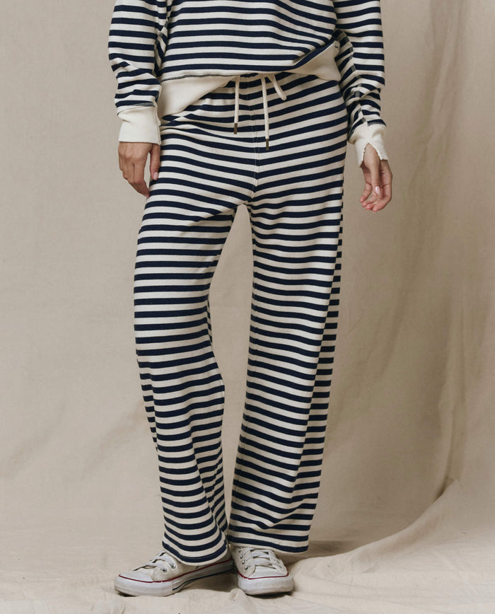 A person in THE GREAT. SP26 SEASONAL CORE The Lap Sweatpant -- Navy Sailor Stripe, paired with a matching navy and white striped long-sleeve top and white sneakers, stands before a beige background.