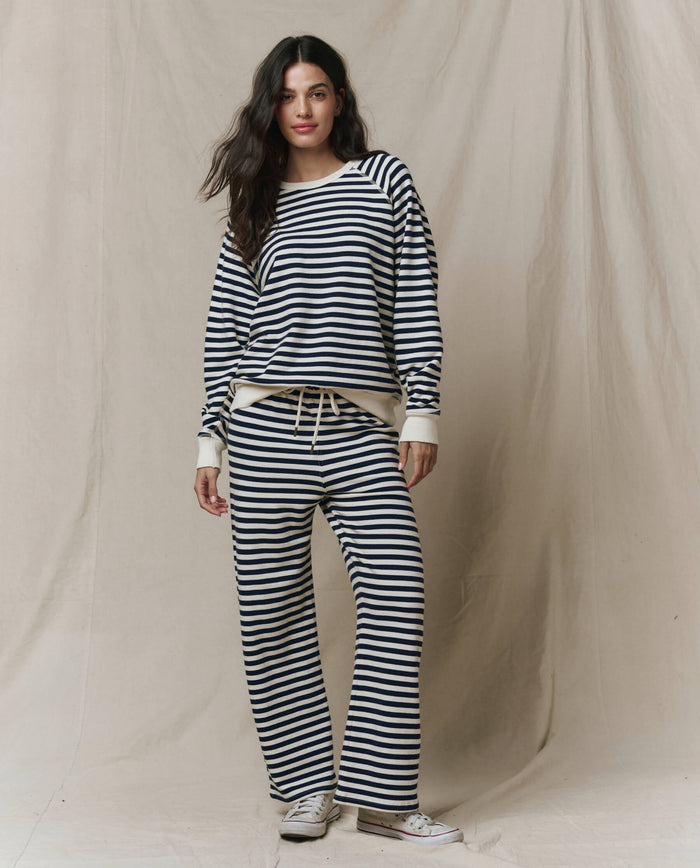 Wearing a matching black and white striped long-sleeve top and THE GREAT. SP26 SEASONAL CORE The Lap Sweatpant in Navy Sailor Stripe with white sneakers, a person stands before a beige fabric backdrop.