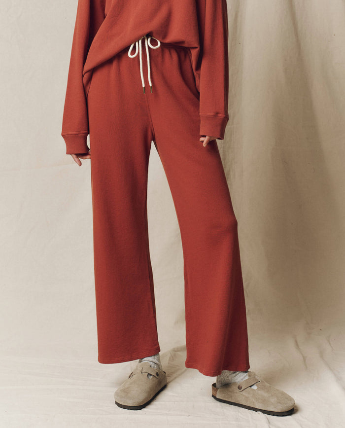A person wears THE GREAT. F25 SEASONAL KNITS’ The Lap Sweatpant, Solid -- Brick—loose-fitting with a drawstring waist—paired with a matching sweater, grey socks, and beige slip-ons against a neutral fabric backdrop.