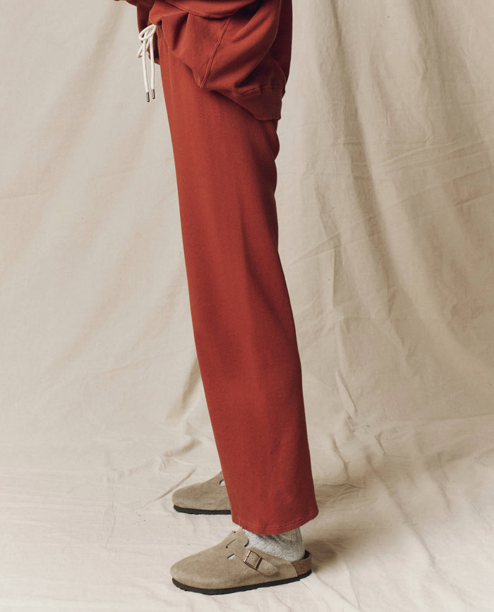 Person wears THE GREAT. F25 SEASONAL KNITS The Lap Sweatpant, Solid -- Brick, with a matching sweatshirt, gray socks, and beige sandals. Wide cropped sweatpants are handcrafted in Los Angeles for casual comfort.