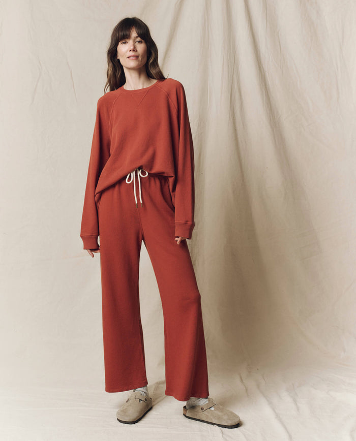 A woman stands against a beige backdrop, wearing a loose rust-colored sweatshirt and THE GREAT. F25 SEASONAL KNITS The Lap Sweatpant in Solid Brick, featuring a wide cropped fit, paired with light-colored shoes.