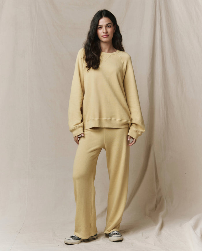 A woman stands before a beige backdrop wearing THE GREAT. SP26 SEASONAL CORE The Lap Sweatpant in Butter Yellow—cropped, loose-fitting, handcrafted in Los Angeles for effortless style—paired with a matching sweatshirt and casual shoes.