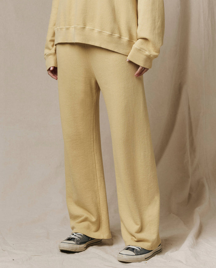 A person wears THE GREAT. SP26 SEASONAL CORE The Lap Sweatpant in Butter Yellow with a matching sweatshirt, standing indoors against a neutral fabric backdrop. Only the lower body is visible; the handcrafted pants feature a cropped, straight-leg fit.