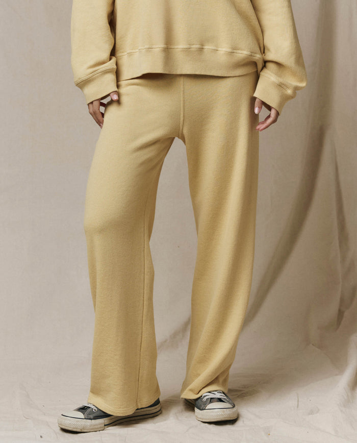 A person in a beige sweatshirt and THE GREAT. SP26 SEASONAL CORE The Lap Sweatpant in Butter Yellow stands on light fabric, hands at their sides, pairing the look with black and white sneakers.