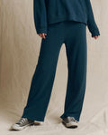 Clad in THE GREAT. F26 SEASONAL CORE’s The Lap Sweatpant. Solid in Dark Slate, paired with a coordinating sweatshirt and black low-top sneakers, a person stands before a beige fabric backdrop, capturing the relaxed spirit of handcrafted Los Angeles style.