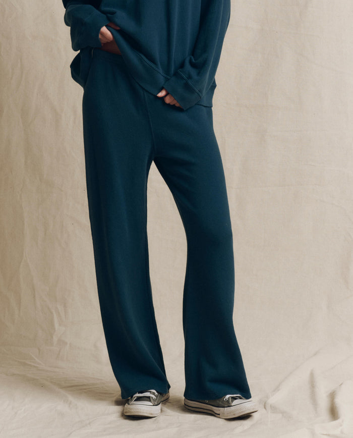 A person wearing loose-fitting, dark slate The Lap Sweatpant by THE GREAT. F26 SEASONAL CORE—wide cropped sweatpants handcrafted in Los Angeles—stands against a beige fabric backdrop with hands tucked in pockets.