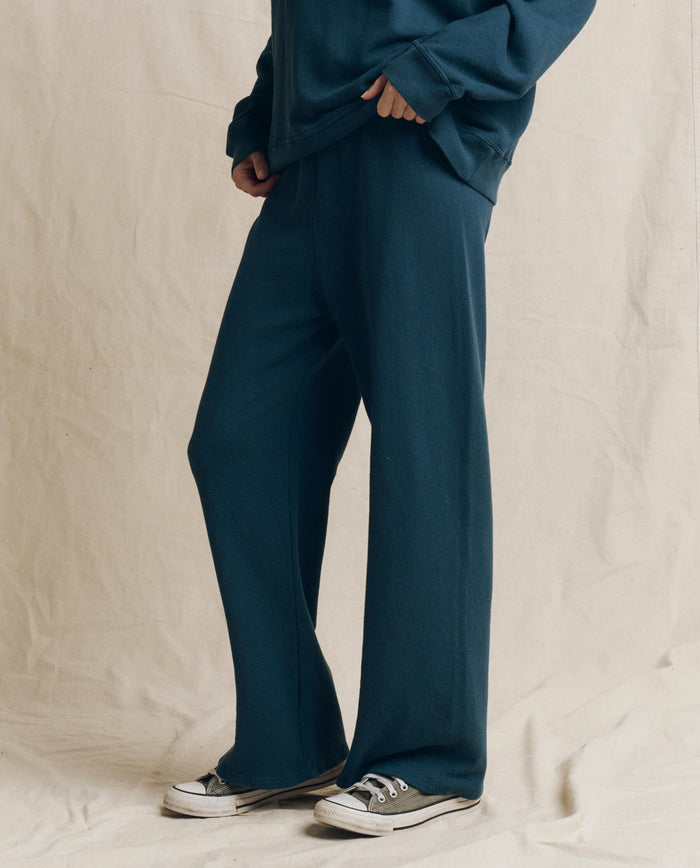 A person wearing THE GREAT. F26 SEASONAL CORE The Lap Sweatpant, Solid -- Dark Slate, along with a matching sweatshirt and gray sneakers, stands against a beige fabric backdrop with only their lower body visible.