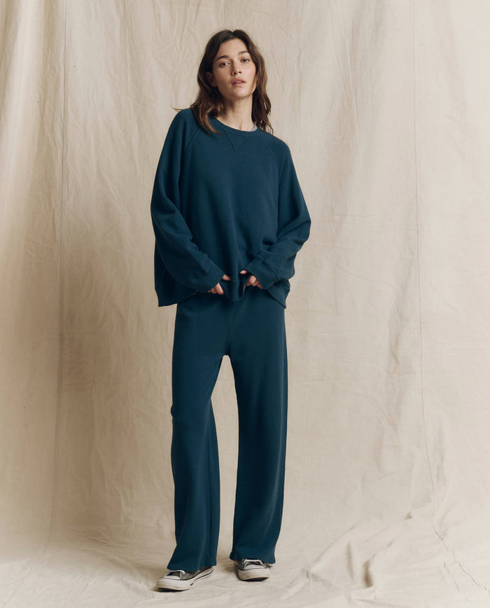 A person stands against a beige fabric backdrop, wearing a loose-fitting teal sweatshirt and THE GREAT. F26 SEASONAL CORE The Lap Sweatpant in Solid — Dark Slate, paired with black-and-white sneakers.